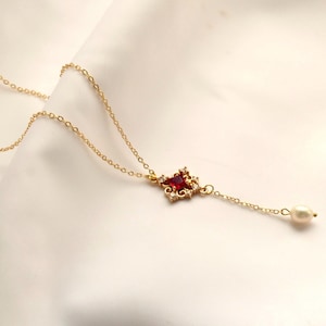 Peut inclure: Un collier à chaîne dorée doté d'un pendentif carré central orné d'une pierre précieuse rouge foncé entourée de petites pierres claires, terminé par une délicate perle blanche suspendue au bout de la chaîne, formant un bijou raffiné.
