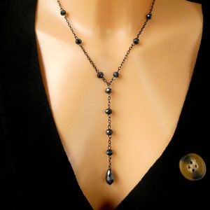 May include: A black beaded Y-shaped necklace with a delicate chain. The necklace features small, dark beads and a teardrop-shaped pendant. The jewelry is displayed on a person wearing a black top.