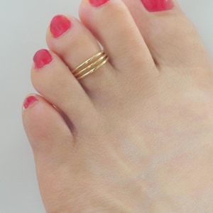 Op de afbeelding: Een goudkleurige teenring met een ontwerp van drie bandjes. De ring bestaat uit drie dunne, gladde, parallelle bandjes die samengevoegd zijn tot één sieraad, gedragen om de tweede teen met felroze nagellak op de nagels.