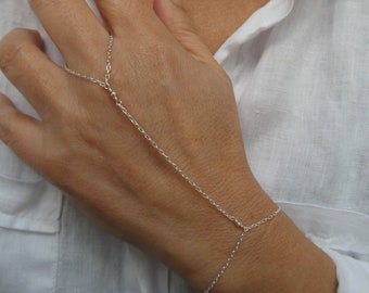 Puede incluir: Un brazalete de cadena de plata con un anillo que se conecta a la muñeca. El brazalete está hecho de una cadena delicada y tiene pequeñas cuentas en los puntos donde la cadena se divide.