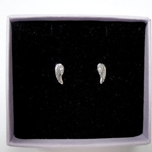 Puede incluir: Un par de pendientes de bot&oacute;n plateados con forma de ala, presentados en una caja forrada de terciopelo negro con borde lavanda. Los pendientes son peque&ntilde;os y detallados, con dise&ntilde;os de plumas.