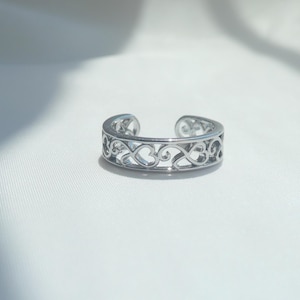 May include: A silver-toned open band ring featuring a continuous cut-out pattern of interconnected heart shapes and swirling filigree details. The ring has a polished metallic finish and a gap at the top, allowing for an adjustable fit on the finger.