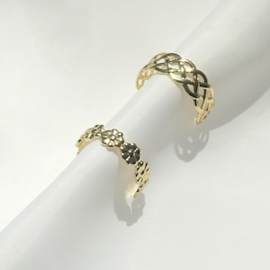 Op de afbeelding: Twee goudkleurige ringen. Eén ring is voorzien van een doorlopende band met kleine, in reliëf aangebrachte bloemvormen. De tweede ring vertoont een ingewikkeld, opengewerkt gevlochten patroon. Beide ringen hebben een gepolijste, metalen afwerking.