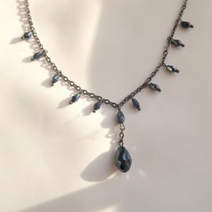 Può includere: Una collana a catena in metallo grigio scuro con una serie di piccole perle di cristallo nero sfaccettato che pendono lungo la catena, terminando con un ciondolo in cristallo nero più grande a forma di goccia al centro della collana.