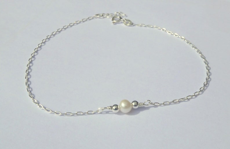 May include: A delicate silver anklet featuring a small, lustrous pearl and two silver beads. The anklet is made of a fine chain and is designed to be worn around the ankle. The pearl is approximately 0.25 inches in diameter.