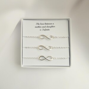 May include: A set of three silver infinity symbol bracelets presented in a white gift box. The box includes the text "The love between a mother and daughter is Infinite". The bracelets are delicate and connected by a chain.