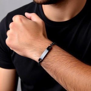 May include: A black bracelet with a rectangular plate and chain links. The bracelet is worn on a wrist, showcasing its sleek, minimalist design. The dark color of the bracelet contrasts with the skin tone, highlighting its modern aesthetic.
