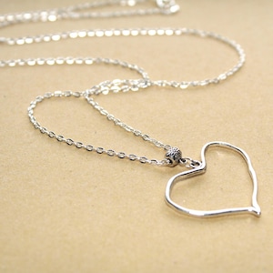 May include: A silver-toned necklace featuring a thin, open-frame heart pendant. The pendant is attached to a delicate cable chain by a small, textured, cylindrical metal bead. The metal has a polished, reflective finish throughout the entire piece.