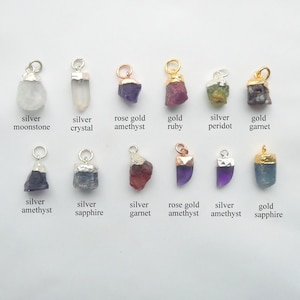 May include: A collection of twelve raw gemstone pendants arranged in two rows, featuring moonstone, crystal, amethyst, ruby, peridot, garnet, and sapphire stones set in silver, gold, and rose gold metal caps with attached circular jump rings.