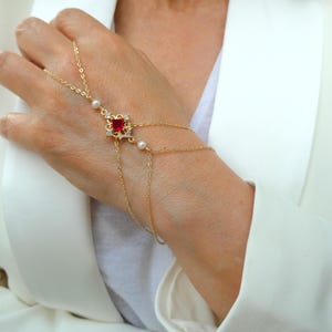 May include: Gold hand chain bracelet featuring a square red gemstone center with surrounding clear stones. The bracelet includes delicate chains and small white pearls. A stylish piece of jewelry.