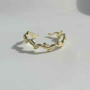Op de afbeelding: Een goudkleurige open ring met een doorlopend rankachtig ontwerp met kleine, gladde, gepolijste blaadjes die om de band heen krullen. De ring heeft een glanzende metalen afwerking en een verstelbare opening aan de bovenkant van het sieraad.