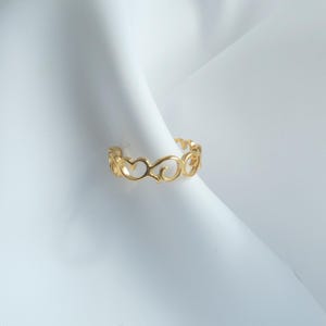 Può includere: Un anello dorato con un delicato design traforato a forma di cuori e volute. L'anello è esposto su uno sfondo di tessuto bianco, evidenziando i suoi intricati dettagli e la calda tonalità metallica. Il design dell'anello è simmetrico ed elegante.