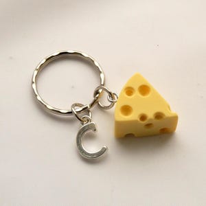 May include: A silver key chain with a letter C charm and a yellow cheese charm. The cheese charm is shaped like a wedge of Swiss cheese with holes.
