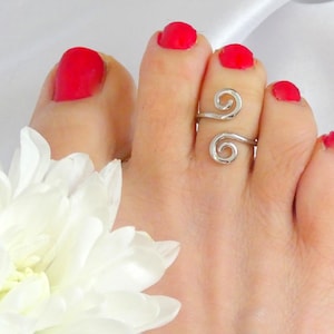 Puede incluir: Un anillo de dedo del pie plateado con un diseño de doble espiral. El anillo se usa en un pie con esmalte de uñas rojo. Una flor blanca está en la esquina inferior izquierda.