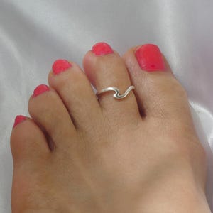 Puede incluir: Un anillo para el dedo del pie de tono plateado con un diseño ondulado suave y continuo. El anillo se lleva en el segundo dedo, que tiene esmalte de uñas rosa brillante. La banda de metal es fina y pulida, creando una silueta curva simple.