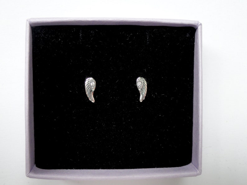 Puede incluir: Un par de pendientes de bot&oacute;n plateados con forma de ala, presentados en una caja forrada de terciopelo negro con borde lavanda. Los pendientes son peque&ntilde;os y detallados, con dise&ntilde;os de plumas.