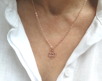 Op de afbeelding: Een delicate roségouden ketting met een kleine, verweven knoop-hanger. De ketting wordt getoond tegen een witte overhemdkraag. De fijne ketting en de gecentreerde hanger creëren een eenvoudige en elegante uitstraling. Een delicaat sieraad.