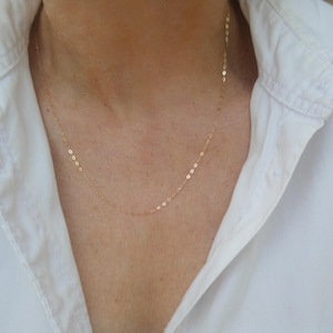 May include: A fine gold necklace with a delicate chain. The necklace is shown against a white collared shirt. The chain is made of small, oval-shaped links. The necklace is a simple, elegant piece of jewellery.