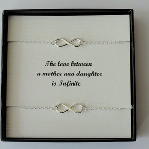 May include: A boxed set of two silver infinity symbol bracelets. The bracelets are presented on a white card with the text "The love between a mother and daughter is Infinite" in black. The box is black.