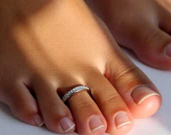 May include: A silver toe ring with small, clear gemstones encircles a toe. The image shows a close-up of a foot with a ring, with other toes visible in the background. The ring is set against a light background.