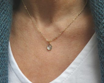 Puede incluir: Un collar de cadena dorada delicada con un pequeño colgante de piedra preciosa translúcida de color verde pálido en forma ovalada. El colgante está sujeto por un engaste dorado sencillo y cuelga de una fina cadena de eslabones dorados pulidos.