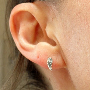 Puede incluir: Un pendiente plateado con forma de ala, con peque&ntilde;as piedras transparentes en el borde. El pendiente se muestra en un piercing en la oreja.