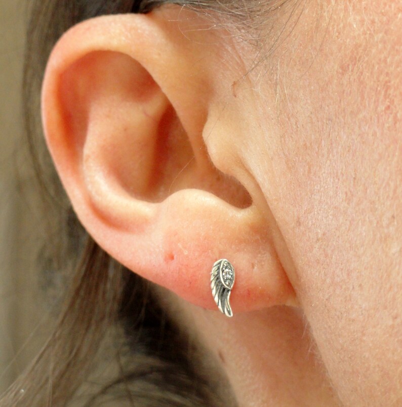 Puede incluir: Un pendiente plateado con forma de ala, con peque&ntilde;as piedras transparentes en el borde. El pendiente se muestra en un piercing en la oreja.