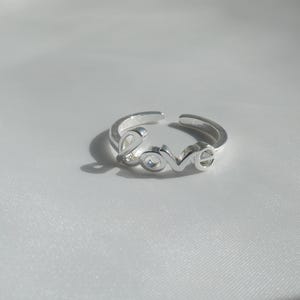 Può includere: Un anello in metallo color argento che forma la parola LOVE in un carattere corsivo stilizzato e collegato. L'anello presenta un design aperto sul retro, che consente di regolare la misura, con una superficie lucida e riflettente.