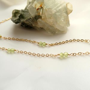 May include: Delicate gold chain necklace with small, light green peridot gemstones. The necklace is displayed on a white surface with a light green crystal formation in the background. A simple and elegant jewelry piece.