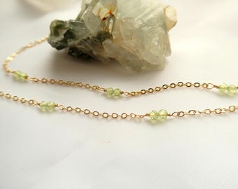 May include: Delicate gold chain necklace with small, light green peridot gemstones. The necklace is displayed on a white surface with a light green crystal formation in the background. A simple and elegant jewelry piece.