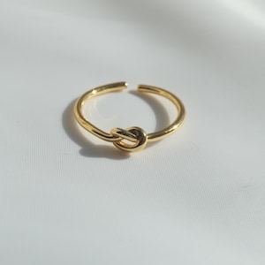 May include: A polished gold-tone ring featuring a central knot design. The band is open at the back, creating an adjustable circular shape with a smooth, reflective metallic finish that catches the light, highlighting the simple knot detail.