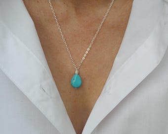 May include: A silver necklace with a teardrop-shaped turquoise pendant. The pendant is a vibrant blue-green color and is suspended from a delicate silver chain. The necklace is worn against a white collared shirt.