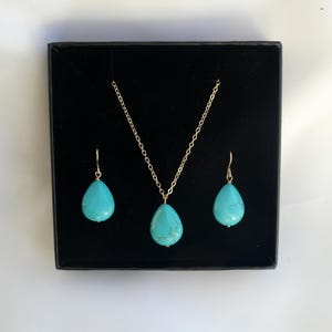 Puede incluir: Un conjunto de joyería que consta de un collar de cadena en tono dorado y dos pendientes a juego, cada uno con una piedra turquesa lisa en forma de lágrima con vetas oscuras, presentado dentro de una caja negra cuadrada con forro aterciopelado.