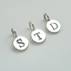 Puede incluir: Tres pequeños dijes circulares de metal plateado, cada uno con una letra mayúscula estampada. Los dijes están dispuestos en diagonal, mostrando las letras S, T y D, con una pequeña argolla de unión fijada en la parte superior de cada pieza.
