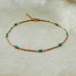 Puede incluir: Una delicada pulsera de cadena dorada con pequeñas cuentas turquesas espaciadas uniformemente. La pulsera se exhibe sobre una tela beige claro, mostrando su elegancia y diseño simples. Es una pieza de joyería.
