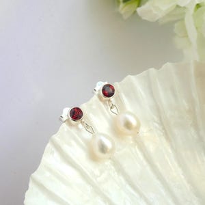 Peut inclure: Boucles d'oreilles en argent avec des pierres rouges et des perles en forme de goutte. Les boucles d'oreilles sont présentées sur une surface blanche texturée, avec un arrière-plan flou.
