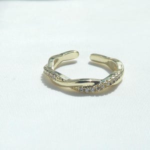 Può includere: Un anello aperto dorato con un design a spirale. L'anello è adornato con piccole pietre preziose trasparenti su un lato, aggiungendo un tocco di brillantezza. L'anello è posto su uno sfondo bianco.