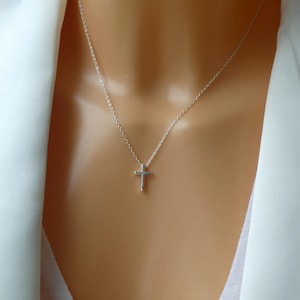 Peut inclure: Un délicat collier avec un pendentif en forme de croix argentée. La croix est petite et suspendue à une fine chaîne. Le collier est présenté sur un fond blanc.