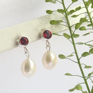 Puede incluir: Un par de pendientes de plata con una piedra roja y una perla blanca colgante en forma de lágrima. Los pendientes se muestran sobre un fondo blanco con follaje verde.