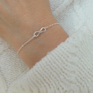 Könnte beinhalten: Ein filigranes Silberarmband mit einem Unendlichkeitszeichen. Das Armband besteht aus einer feinen Kette und wird am Handgelenk getragen, vor dem Hintergrund eines weißen Strickpullovers. Das Unendlichkeitszeichen ist ein klassisches Design.