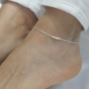 May include: A delicate silver anklet featuring a series of small, white pearls. The anklet is worn on the ankle, showcasing the minimalist design. The chain is thin and the pearls are evenly spaced.