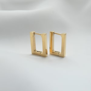 May include: A pair of gold-toned hoop earrings featuring a rectangular, geometric shape with a smooth, polished finish. Each earring is constructed with a hinged post mechanism that secures the rectangular frame, creating a clean, structured look.