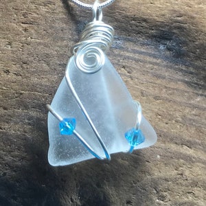Op de afbeelding: Een zilveren draadgewikkelde zeeglas hanger met twee lichtblauwe kristal accenten. De hanger is aan een zilveren ketting.
