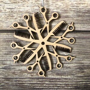 May include: A wooden snowflake ornament with a design of scissors and combs. The ornament is a light brown color and has a rustic look.