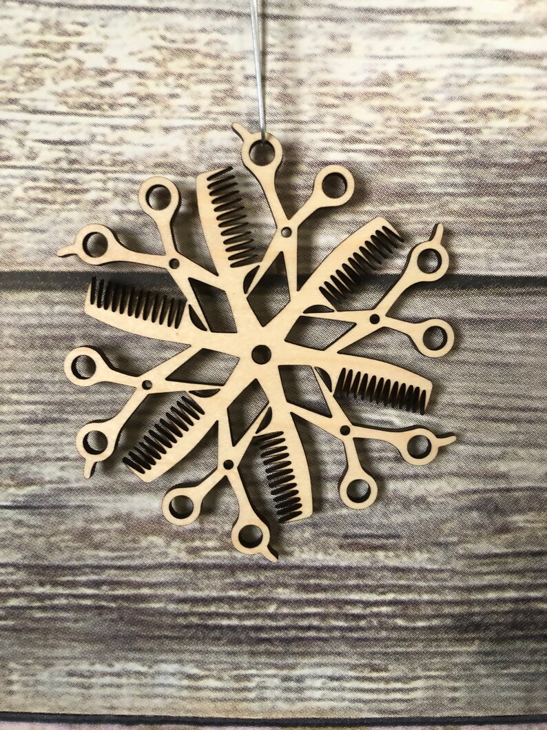 May include: A wooden snowflake ornament with a design of scissors and combs. The ornament is a light brown color and has a rustic look.