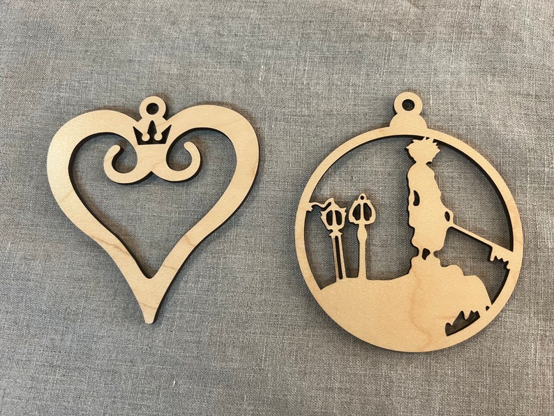 May include: Two wooden ornaments, one shaped like a heart with a crown and the other a circle with a silhouette of a person holding a keyblade.