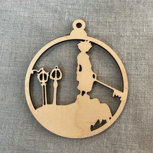 May include: A wooden ornament shaped like a circle with a silhouette of a person holding a keyblade and two keyholes. The ornament is a light brown color.