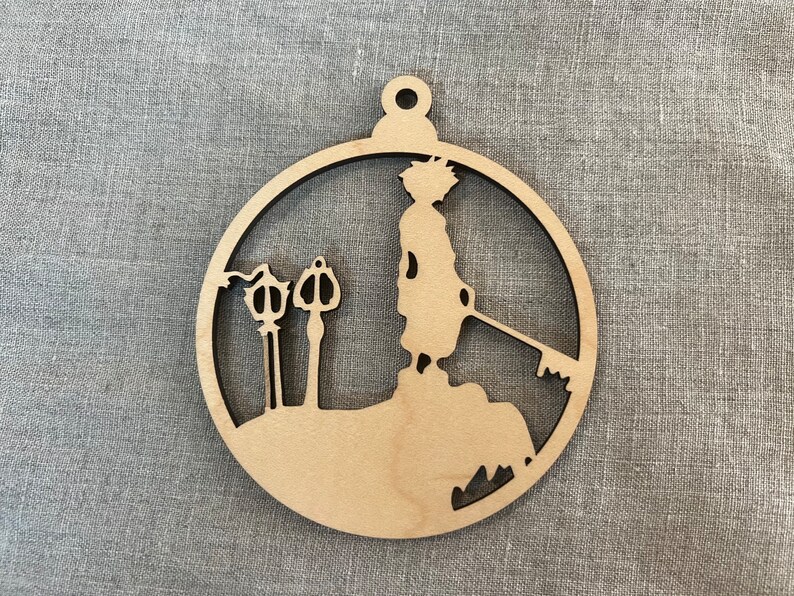 May include: A wooden ornament shaped like a circle with a silhouette of a person holding a keyblade and two keyholes. The ornament is a light brown color.