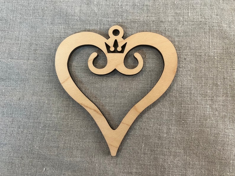 May include: A wooden heart-shaped ornament with a crown design in the center. The ornament is made of light brown wood and has a smooth finish.
