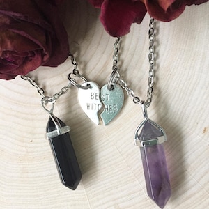May include: Two necklaces with silver chains and pendants. One pendant is a black crystal point, the other is a purple crystal point. The two pendants are connected by a silver heart charm that says "Best Witches" in two halves.